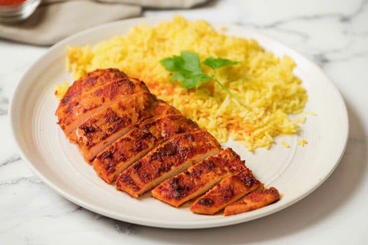 Horizontal shot - Smoked Paprika Chicken Breast with boneless skinless breast sliced, saffron rice garnished with cilantro in background, on a white plate with white marble background.