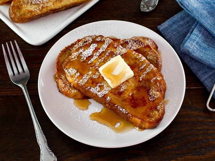 Overhead shot of Challah French Toast with butter, powdered sugar and syrup on a white plate with fork, placed on wooden table with cloth napkin.