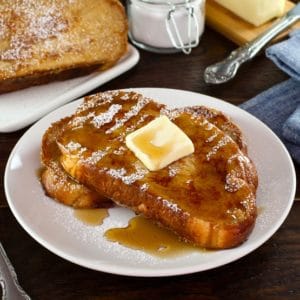 Front angle shot of Challah French Toast with butter, powdered sugar and maple syrup on a white plate with fork, placed on wooden table with cloth napkin. Powdered sugar and plate of French Toast in background.