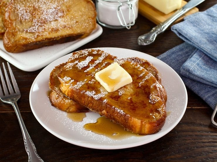 Front angle shot of Challah French Toast with butter, powdered sugar and maple syrup on a white plate with fork, placed on wooden table with cloth napkin. Powdered sugar and plate of French Toast in background.