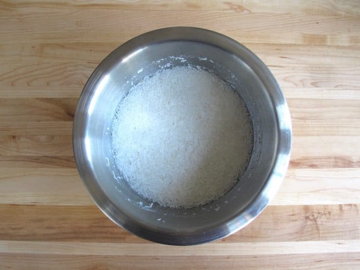Coconut shreds covered with water, soaking in stainless steel mixing bowl on wooden background.