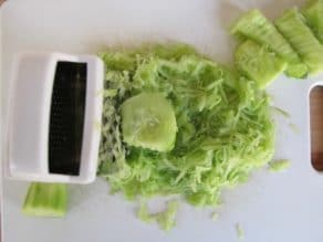 Peeled grated cucumbers with shreds and white grater on a white cutting board.