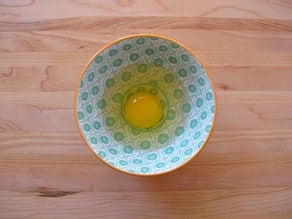 Single egg cracked, uncooked, in small decorative bowl on wooden cutting board.