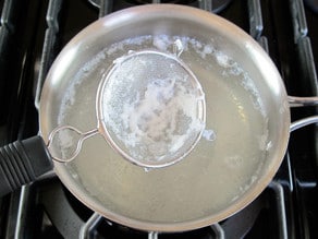 Mesh strainer collecting strands of egg from saucepan with simmering water on stovetop.