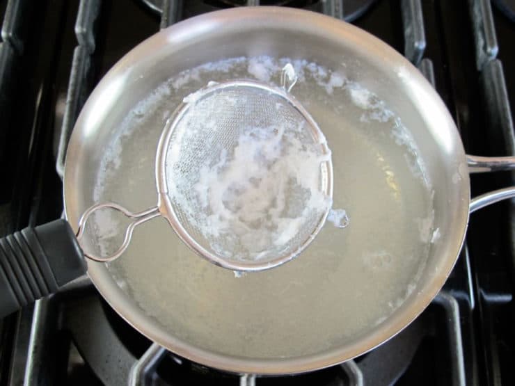 Mesh strainer collecting strands of egg from saucepan with simmering water on stovetop.