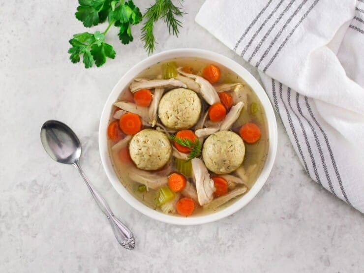 Overhead shot of Jewish matzo ball soup wtih three fluffy matzo balls, chicken, carrot slices and celery pieces in golden broth. Bowl rests on a white marble countertop with spoon, linen towel, and fresh herbs on the side.