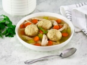 Close up shot of matzo ball soup - classic Jewish chicken soup with fluffy matzo balls on a white countertop. Spoon and linen napkin beside the bowl, white ceramic jar in background.