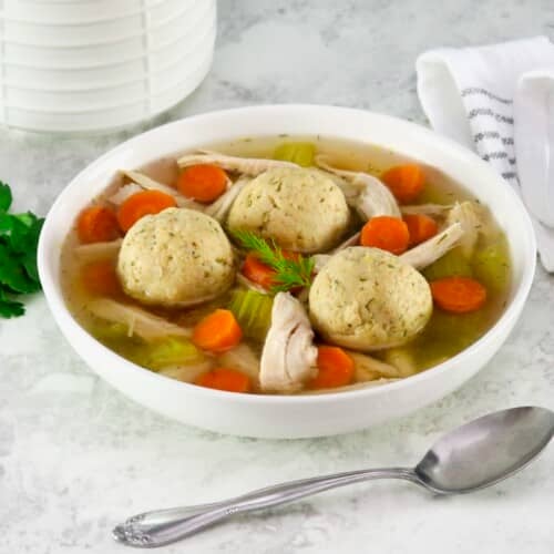 Close up shot of matzo ball soup - classic Jewish chicken soup with fluffy matzo balls on a white countertop. Spoon and linen napkin beside the bowl, white ceramic jar in background.