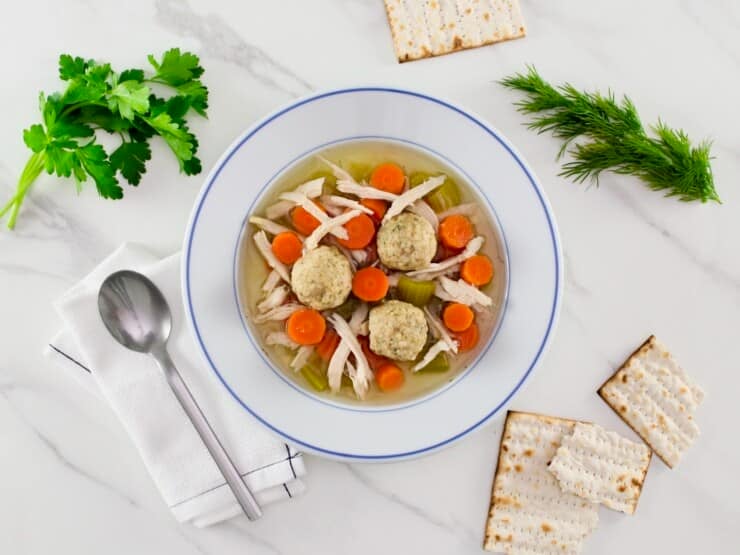 Overhead shot - sinker matzo ball soup on a white marble surface. Soup includes dense sinker matzo balls, chicken, carrots, celery, and golden broth in a white soup bowl with spoon, cloth napkin, cracked matzo and fresh green herbs beside the bowl. Classic Jewish soup.