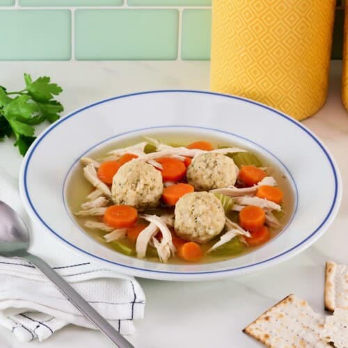 Floater matzo balls in a shallow bowl of Jewish chicken soup with carrot slices, pieces of celery, and golden broth. A spoon, fresh herbs, three small sheets matzo and two yellow large canisters in background.
