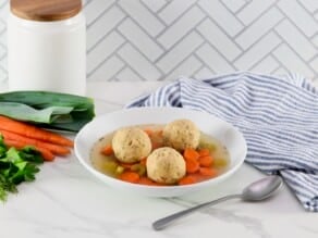 Horizontal shot of fluffy floater matzo balls in a shallow bowl of Jewish vegetarian matzo ball soup with carrots, celery, and golden saffron broth. Bowl rests on a white marble countertop with a spoon, blue striped linen napkin, white jar and fresh vegetables beside it. White tiles in background.