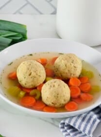 Horizontal shot of floater matzo balls in a shallow bowl of vegetarian matzo ball soup with carrot slices, pieces of celery, and golden saffron broth. Spoon, fresh vegetables, and linen napkin on the white marble counter beside the bowl. Tiles and white jar in background.