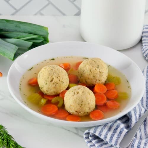 Horizontal shot of floater matzo balls in a shallow bowl of vegetarian matzo ball soup with carrot slices, pieces of celery, and golden saffron broth. Spoon, fresh vegetables, and linen napkin on the white marble counter beside the bowl. Tiles and white jar in background.