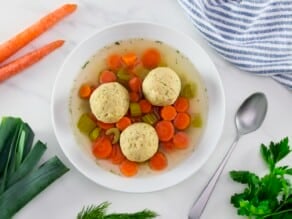 Overhead shot of a bowl of vegetarian matzo ball soup with light and fluffy matzo balls in a shallow bowl with carrot slices, pieces of celery, and golden broth. Spoon, fresh vegetables, and linen napkin on the white marble counter beside the bowl.