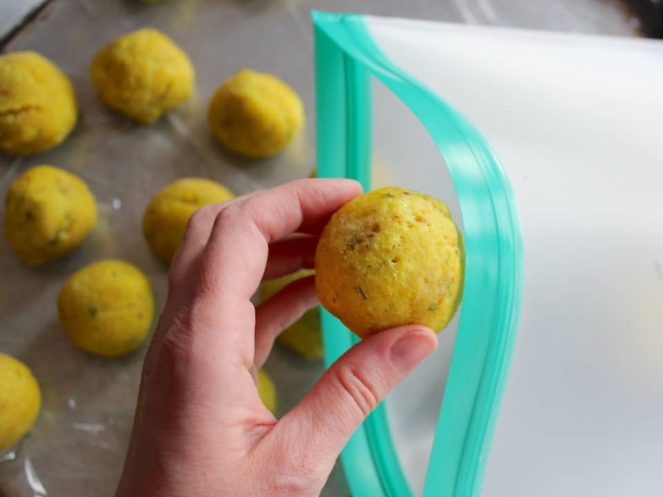Left hand putting a frozen matzo ball into a silicone freezer bag, tray with frozen matzo balls in background.