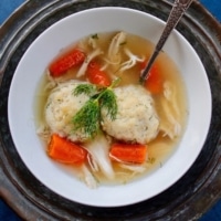 A bowl of matzo ball soup with spoon on a metal platter on blue countertop.