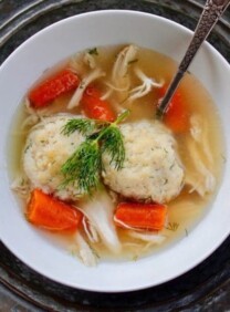A bowl of matzo ball soup with spoon on a metal platter on blue countertop.