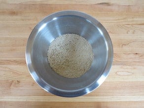 Stainless steel mixing bowl filled with dry matzo meal and seasonings on a wooden cutting board.