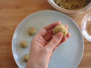 Hand forming matzo ball over a plate of uncooked matzo balls. Mixing bowl of matzo ball batter in the background.