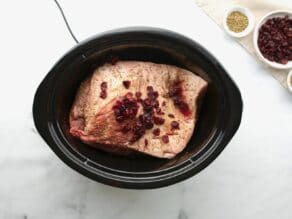Uncooked brisket with cranberries and wine in a slow cooker crock pot with garnishes on the side.