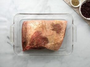 Brisket in glass dish with spices to marinate coating the exterior, garnishes on the side.