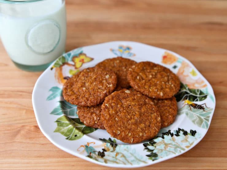 Anzac biscuits arranged on a plate with a beautiful floral design, accompanied by a glass of milk on the left.