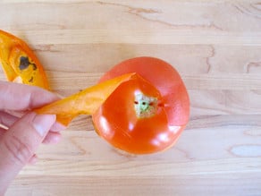 Fingers peeling skin from tomato on cutting board.