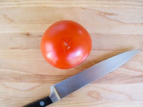Whole tomato and chef's knife on cutting board, X sliced into bottom of tomato.