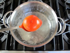 Spoon holding tomato over pot of boiling water on stovetop.