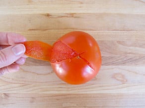 Fingers peeling skin from tomato with X sliced into bottom on cutting board.