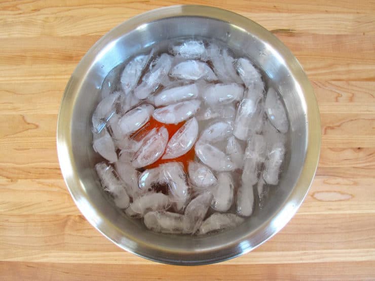 Tomato in bowl of ice water on cutting board.