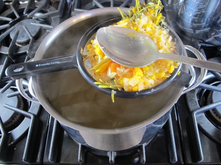 Straining herbs and vegetables from pot of court bouillon, spoon pushing out liquid from the solids in strainer, over pot on stovetop.