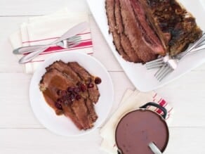 Overhead shot of a white wooden table set with dinner plate of Cranberry Chipotle brisket slices slathered in cranberry sauce and juices, with cloth napkin and utensils. Platter of sliced brisket with two forks rests alongside a golden pan of rich red sauce with a ladle, resting on a cloth napkin.