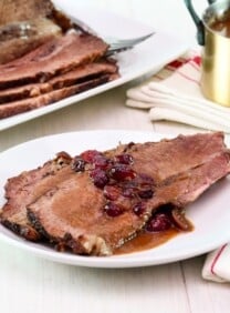 Horizontal shot - white wooden table set with dinner plate of Cranberry Chipotle brisket slices slathered in cranberry sauce and juices, with cloth napkin and utensils. Platter of sliced brisket and golden pan of sauce on a cloth napkin in the background.