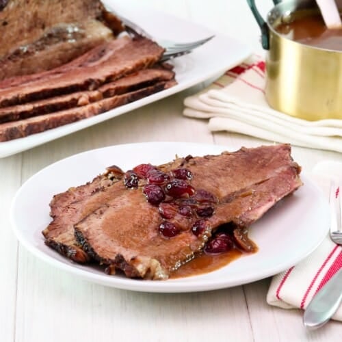 Horizontal shot - white wooden table set with dinner plate of Cranberry Chipotle brisket slices slathered in cranberry sauce and juices, with cloth napkin and utensils. Platter of sliced brisket and golden pan of sauce on a cloth napkin in the background.