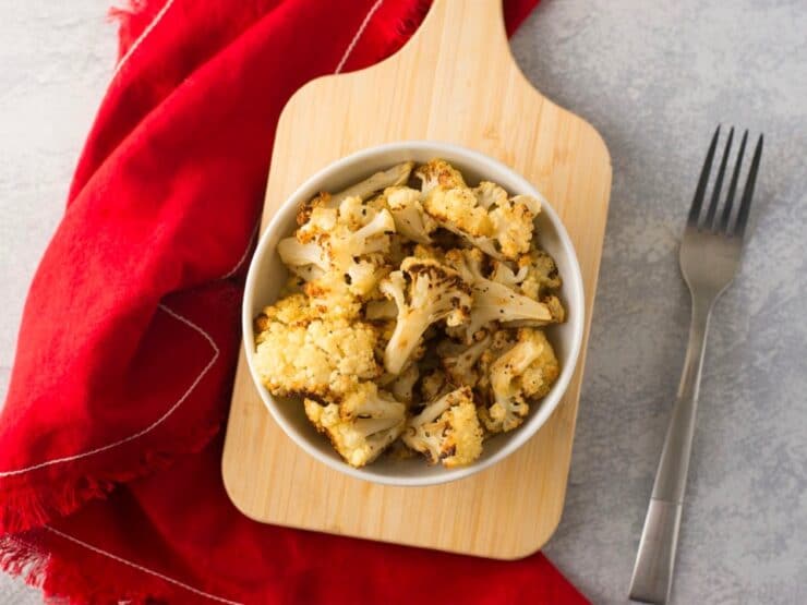 Overhead shot of roasted cauliflower in white dish on wooden cutting board with red napkin and fork in background on a concrete surface.