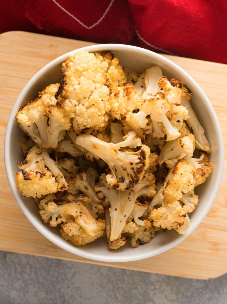 Vertical overhead shot of roasted cauliflower florets in white bowl on small wood cutting board resting on concrete surface, red napkin laying nearby.