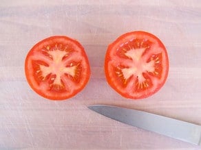 Overhead shot of tomato sliced in half with chef's knife on white plastic cutting board.