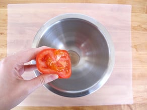 Overhead shot of stainless steel bowl, hand squeezing tomato half into the bowl.