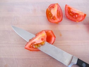 Chef's knife cutting seed pulp away from tomato quarter.