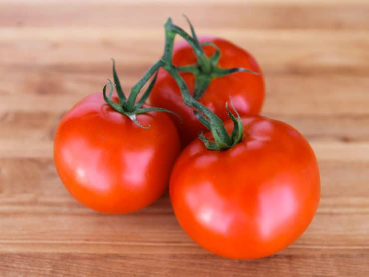 Three whole ripe red tomatoes on the vine, resting on wooden cutting board.