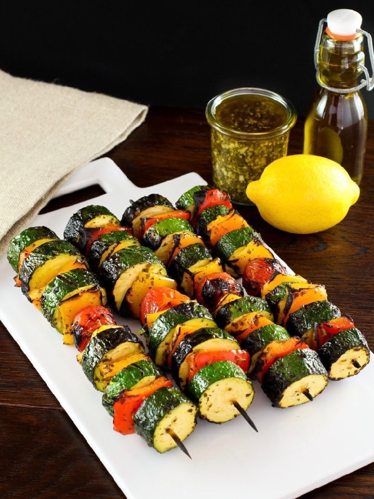 Overhead shot of four lemon pesto vegetable skewers on white plate on wooden table, lemon, pesto and oil in background with napkin.