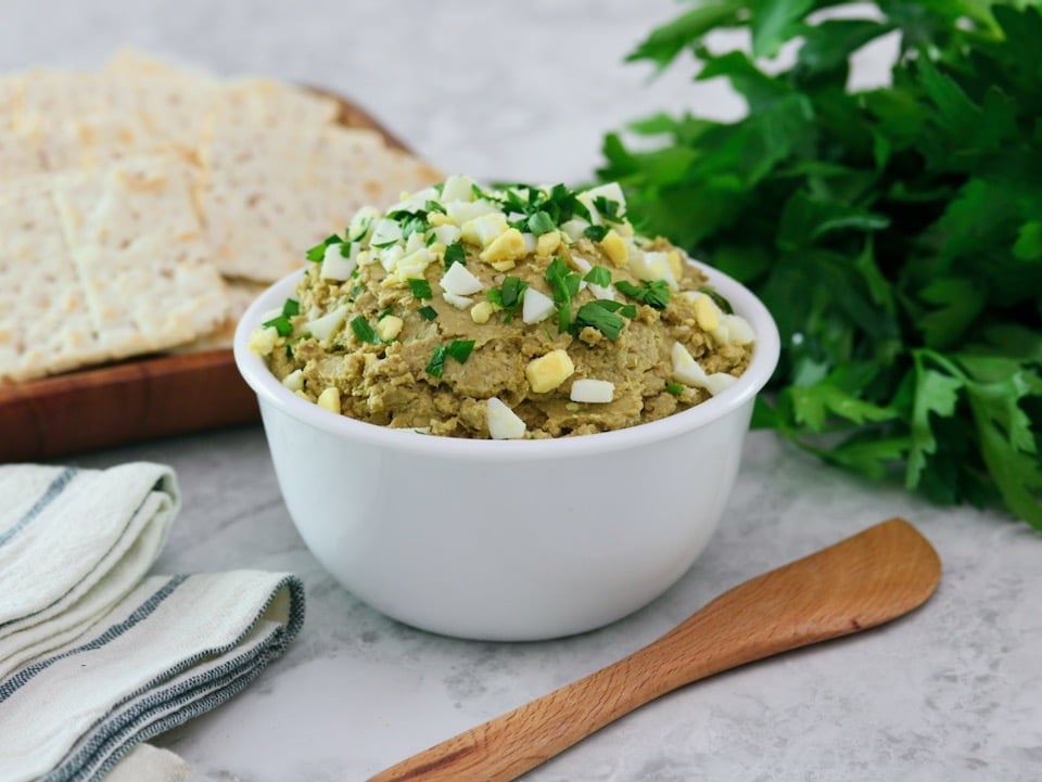 Horizontal shot - White bowl of vegetarian chopped liver topped with chopped hard boiled egg and chopped parsley, with a wooden spatula, bunch of fresh parsley, wooden platter of crackers, and linen napkins beside it, on a marble countertop.