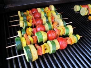 Four lemon pesto vegetable skewers grilling on smoky outdoor barbecue grill.