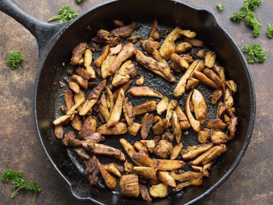 Horizontal image - Cast iron pan with crispy, juicy cooked chicken shawarma in olive oil. Pan is sitting on a brown countertop with fresh parsley scattered around.