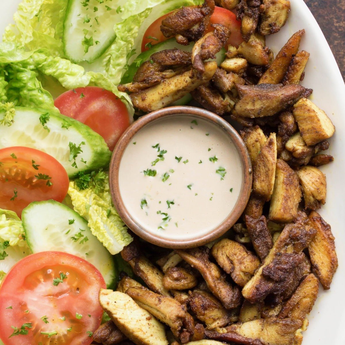 Square crop featured image - white platter of chicken shawarma with fresh lettuce and tomato, small dish of tahini in center garnished with sprinkle of parsley
