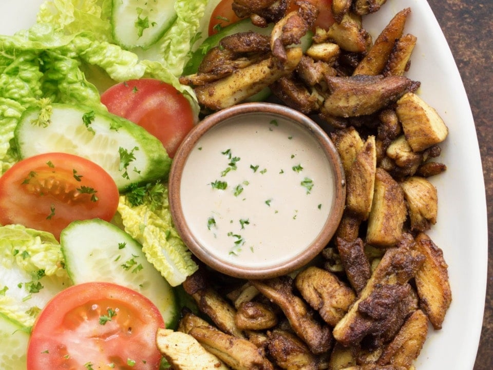 Horizontal close up image - white platter of chicken shawarma with fresh lettuce and tomato, small dish of tahini in center garnished with sprinkle of parsley