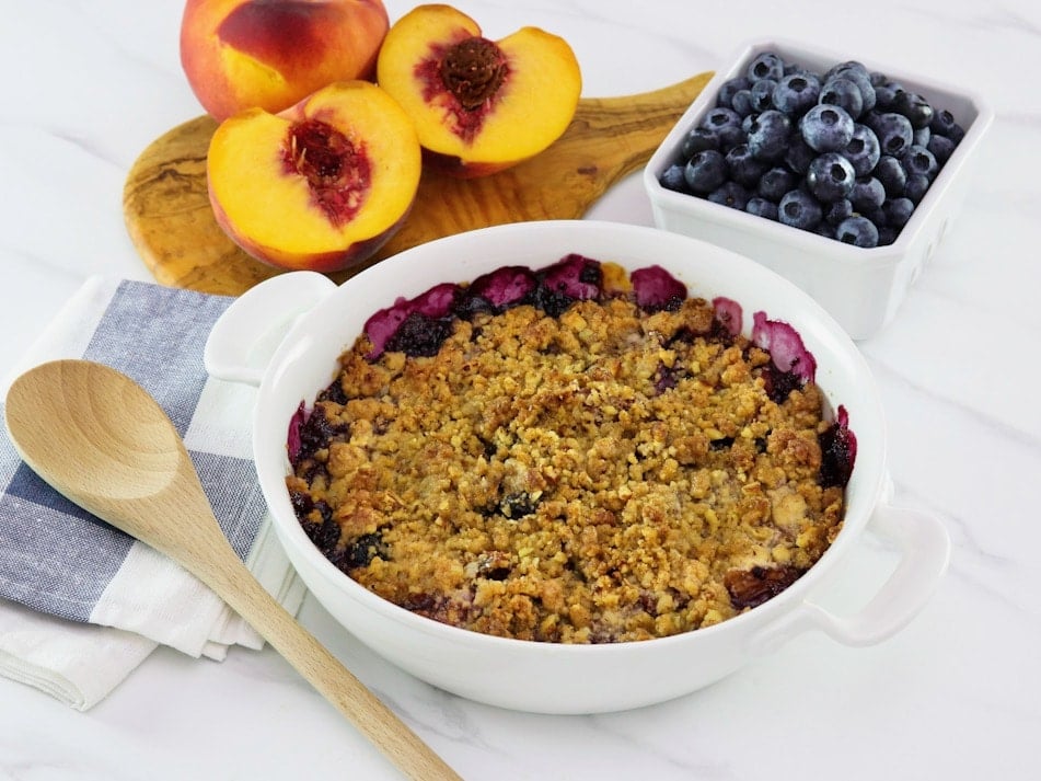 Horizontal image - peach and blueberry crisp in a white baking dish. A wooden spoon is off to the left side and fresh peaches and blueberries are in the background.