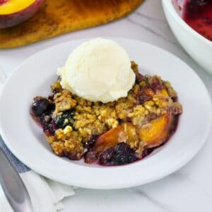 Close-up image - a dish of peach and blueberry crisp topped with a scoop of vanilla ice cream.