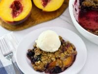 Square image - peach and blueberry crisp on a small white plate, topped with a scoop of vanilla ice cream. A white baking dish of the remaining crisp sits off to the right next to a pile of fresh peaches.